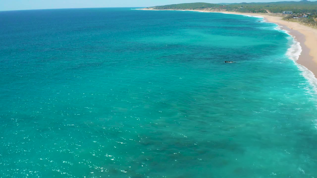 Wide aerial view at El Cardoncito showing bright blue ocean across the foreground, a pale beach at the upper right, and no visible people.