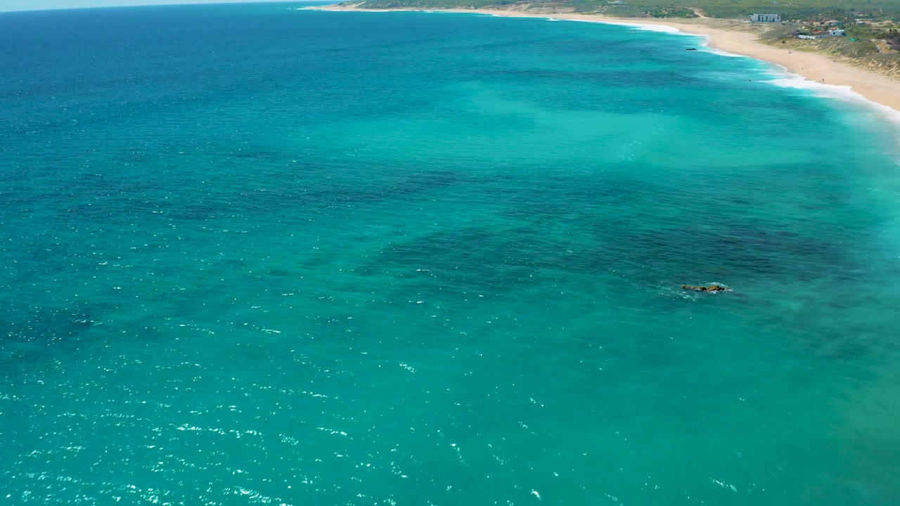 Aerial view over turquoise water at El Cardoncito, Baja California Sur, with a pale beach and low greenery along the upper right, no visible people.