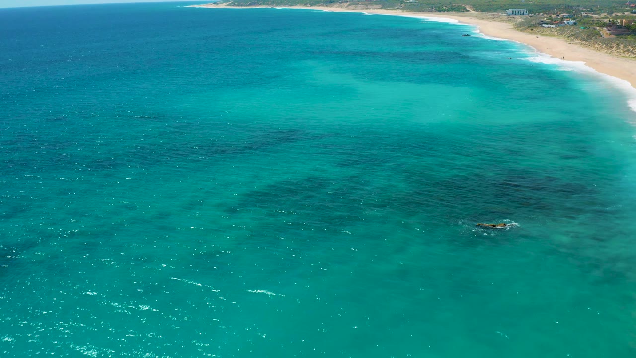 Teal ocean stretches toward the El Cardoncito shoreline, with a broad pale sand strip, low green land behind it, and no visible people in the frame.