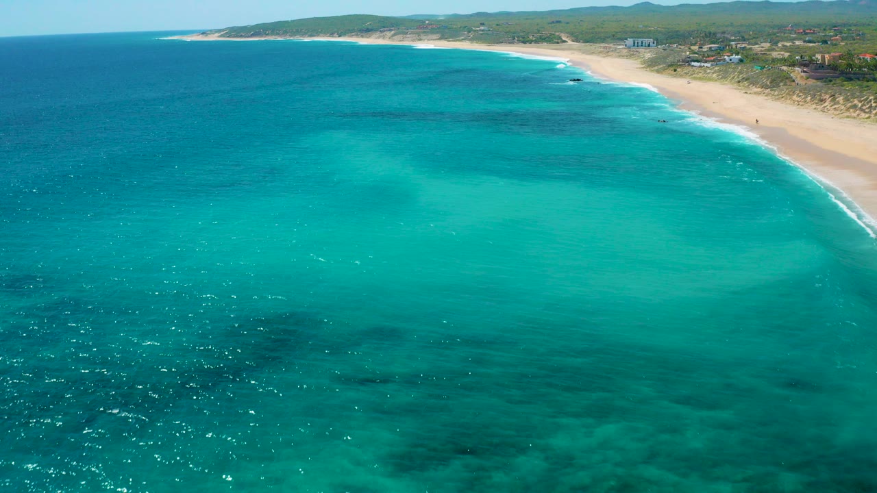 Aerial frame over El Cardoncito, Baja California Sur with turquoise water in the foreground, a curving sandy beach on the right, low inland hills on the horizon, and no visible people.