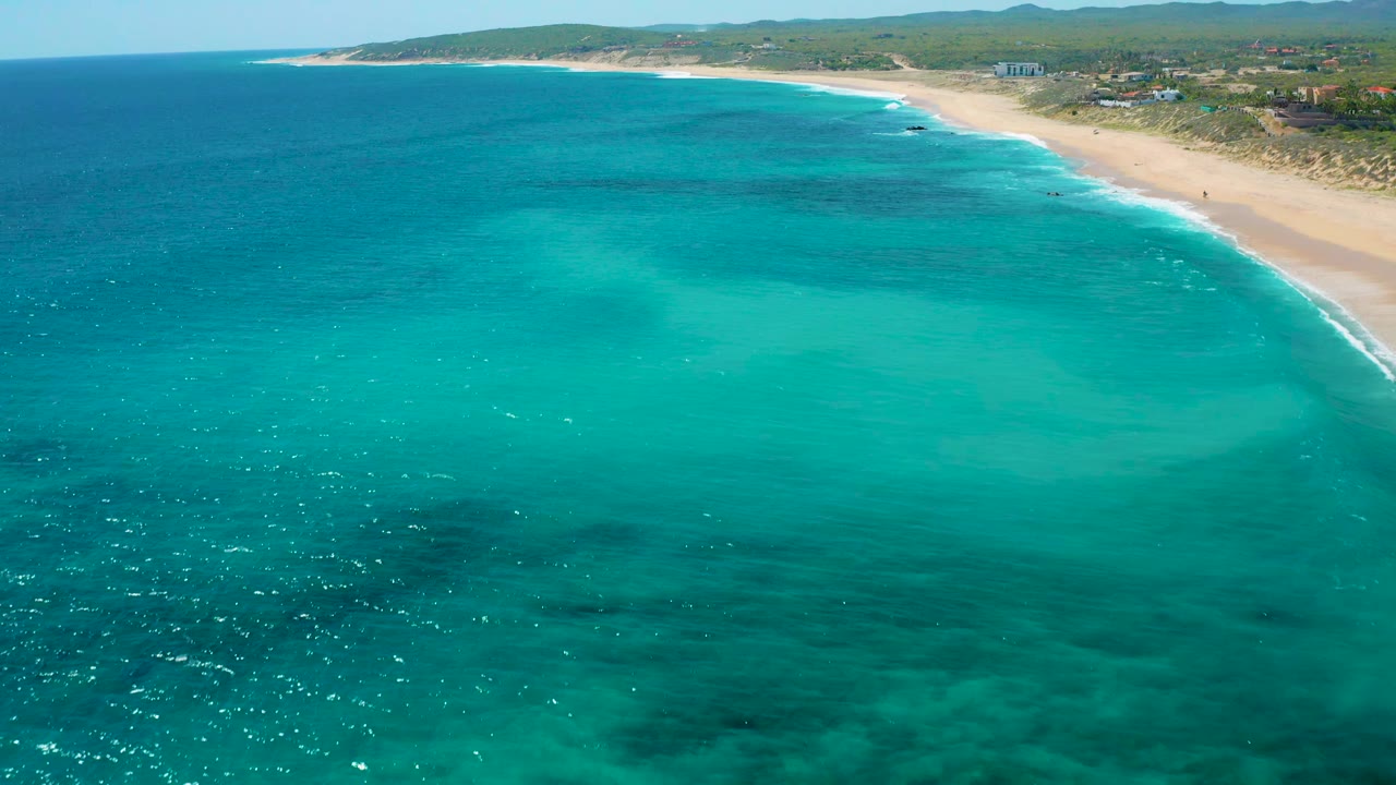 Aerial view in El Cardoncito, Baja California Sur showing blue-green ocean across the foreground, diagonal sand shoreline, small surf breaks, inland vegetation, and no visible people.