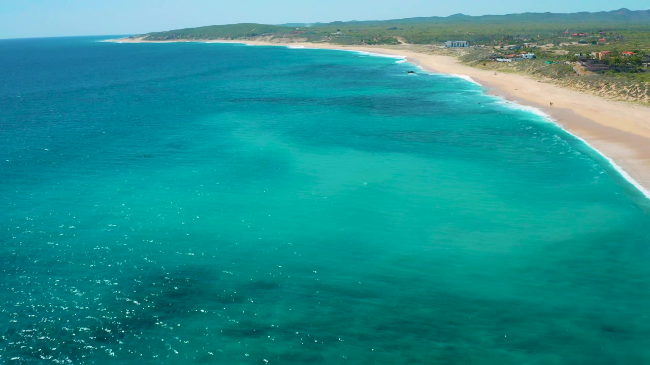 El Cardoncito aerial frame with broad turquoise water, darker underwater patches, pale sand along the right edge, low green scrub inland, scattered structures, and no visible people.