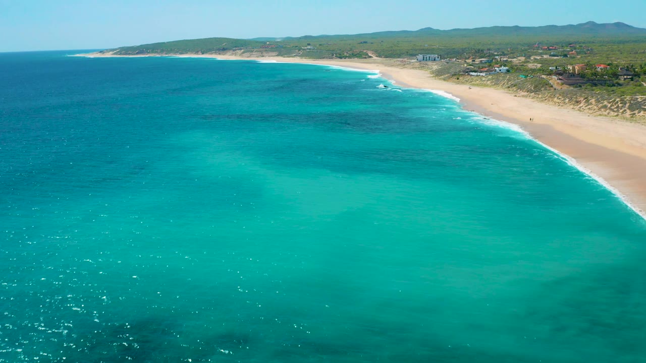 Drone-like view of a sandy beach curving along turquoise ocean under bright sun.