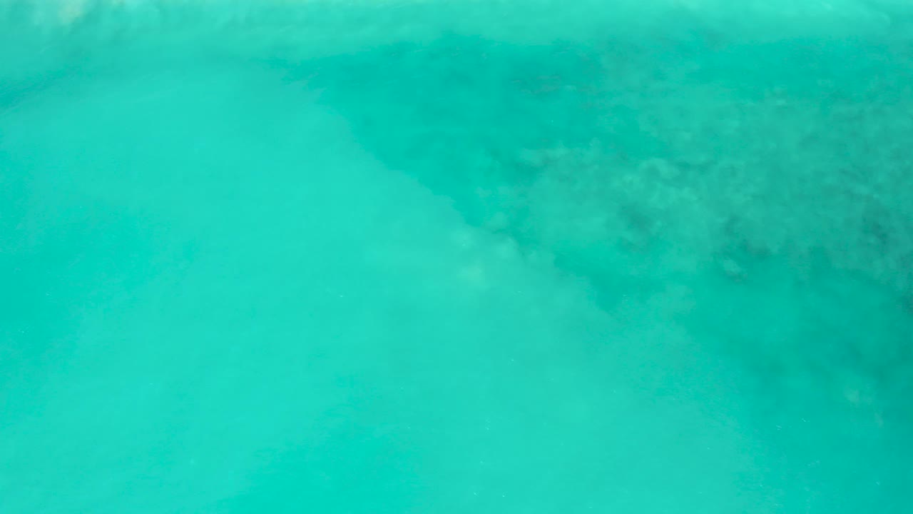 Overhead view of clear turquoise water at El Cardoncito, Baja California Sur, with dark submerged rock or reef shapes near the top and no visible people.
