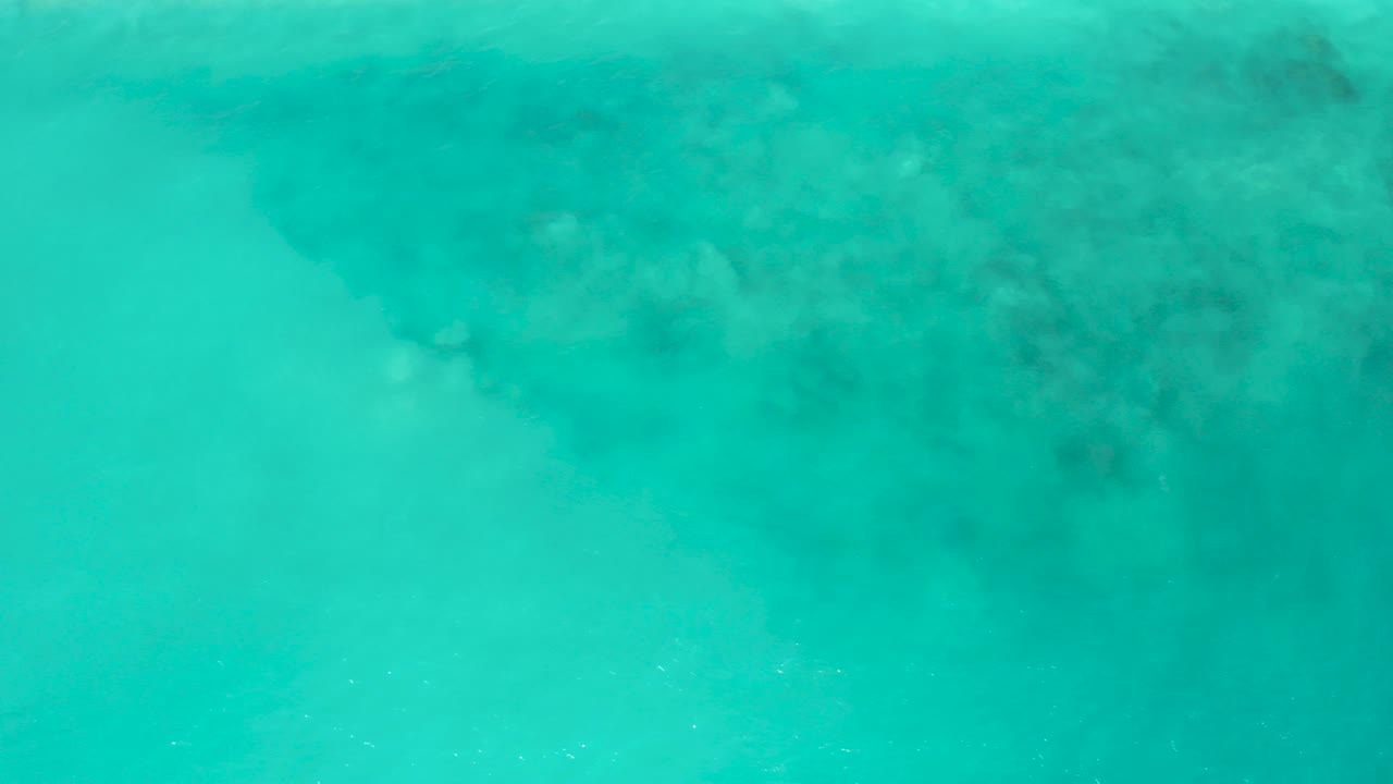 Overhead view of clear turquoise water at El Cardoncito, Baja California Sur, with a darker submerged rock patch near the upper frame and no visible people.