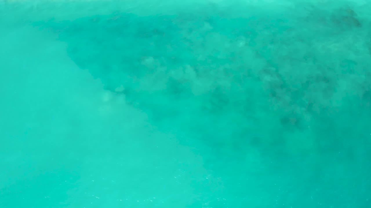 Clear blue-green water fills the frame at El Cardoncito, with a broad dark underwater formation across the upper portion and no visible people.
