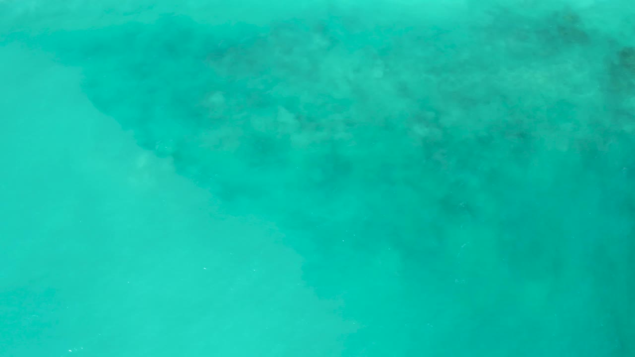 Transparent aqua water at El Cardoncito reveals a darker seafloor mass entering from the upper left, with open pale water below and no visible people.
