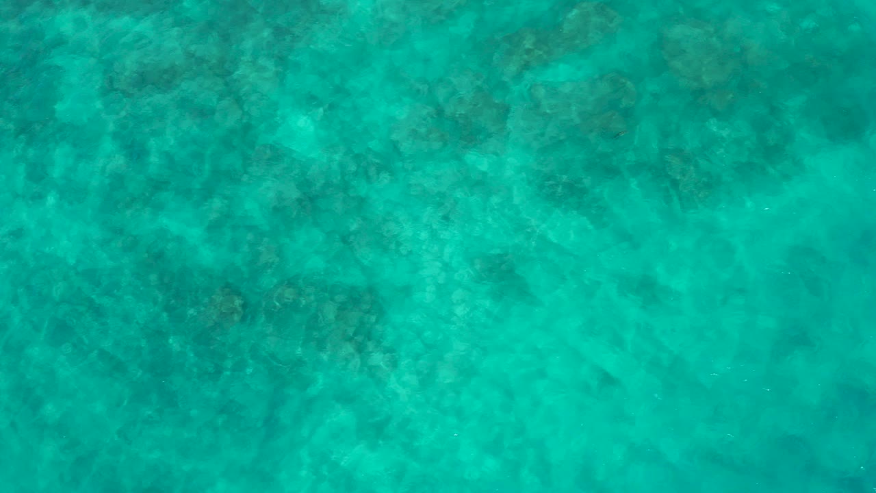 Aerial view of turquoise sea water at El Cardoncito, Baja California Sur, with darker submerged reef or rock texture and no visible people.