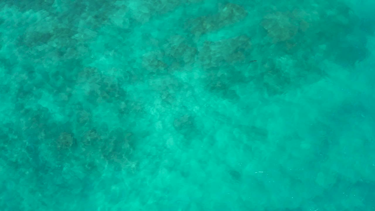Overhead view of shallow-looking turquoise water in Baja California Sur, with pale wave streaks crossing darker underwater shapes and no visible people.