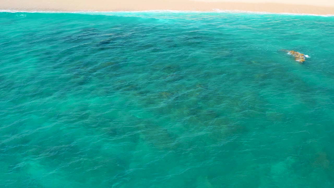 Aerial view over turquoise and deep green water at El Cardoncito, Baja California Sur, with pale beach at the top edge, submerged dark patches, and no visible people.