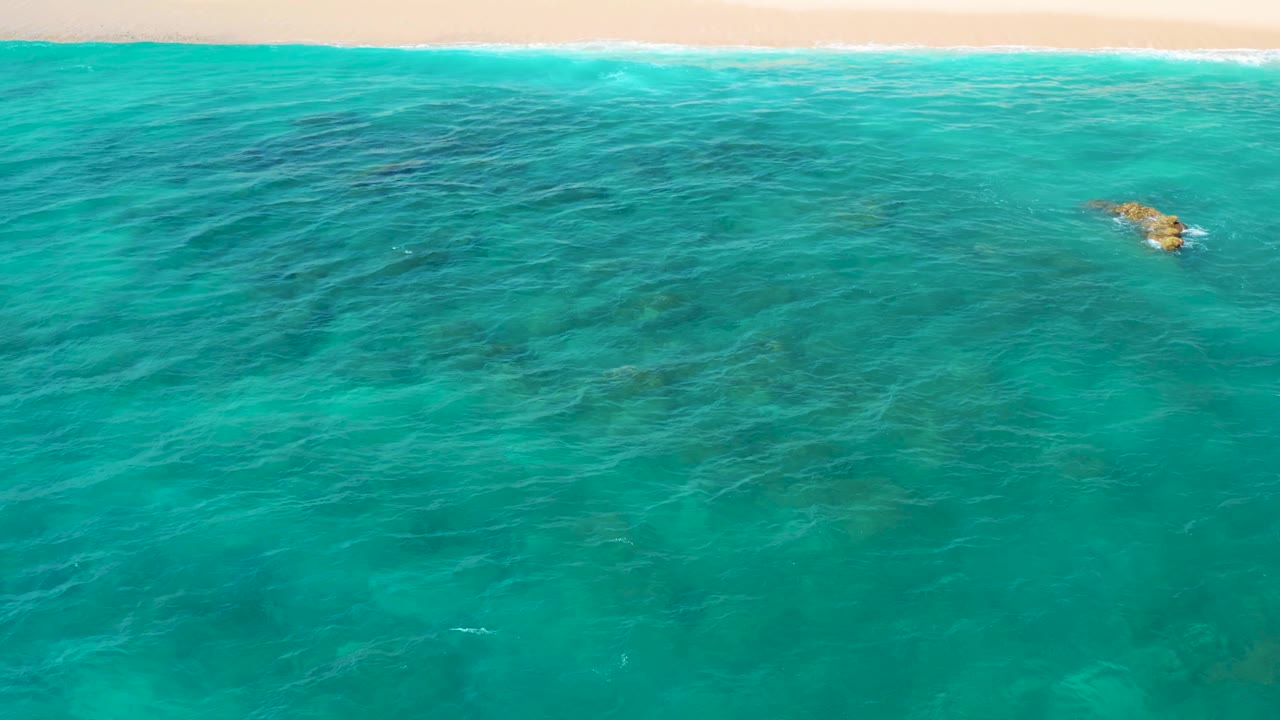 Aerial frame from El Cardoncito showing green-blue ocean, pale beach and white surf across the top, a small reef or rock patch near the right side, and no visible people.