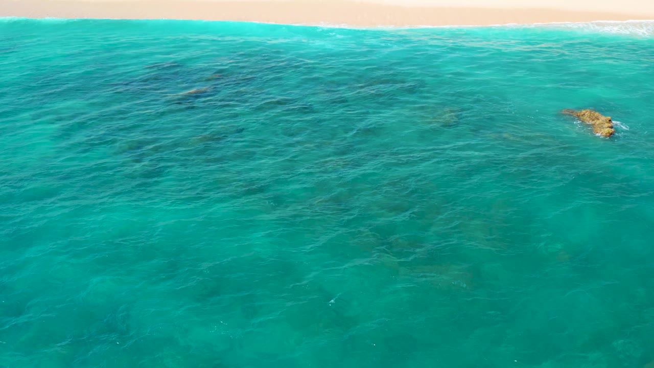 Drone-like overhead view at El Cardoncito with turquoise water in the foreground, a pale sandy shoreline and breaking foam at the top, darker submerged patches, and no visible people.