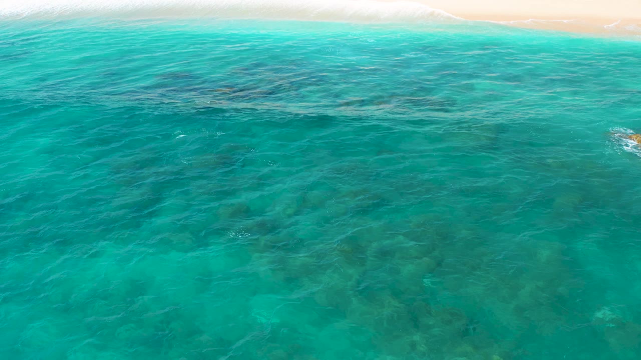 Clear blue-green water covers scattered dark rock patches while a thin strip of sand and shore break sits at the top of the El Cardoncito frame, with no visible people.