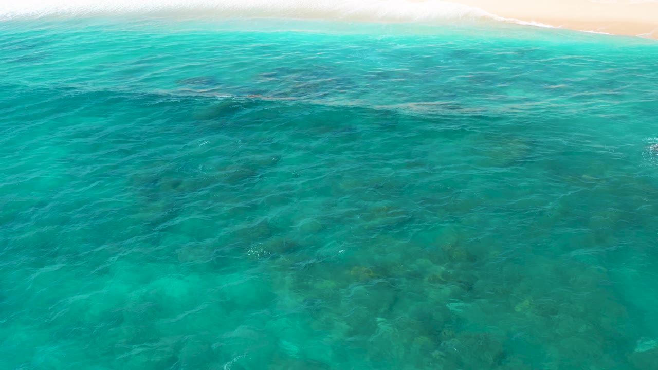 The aerial composition looks down across shallow turquoise sea at El Cardoncito, where darker underwater rocks sit below soft surface ripples and the beach remains along the top edge, with no visible people.