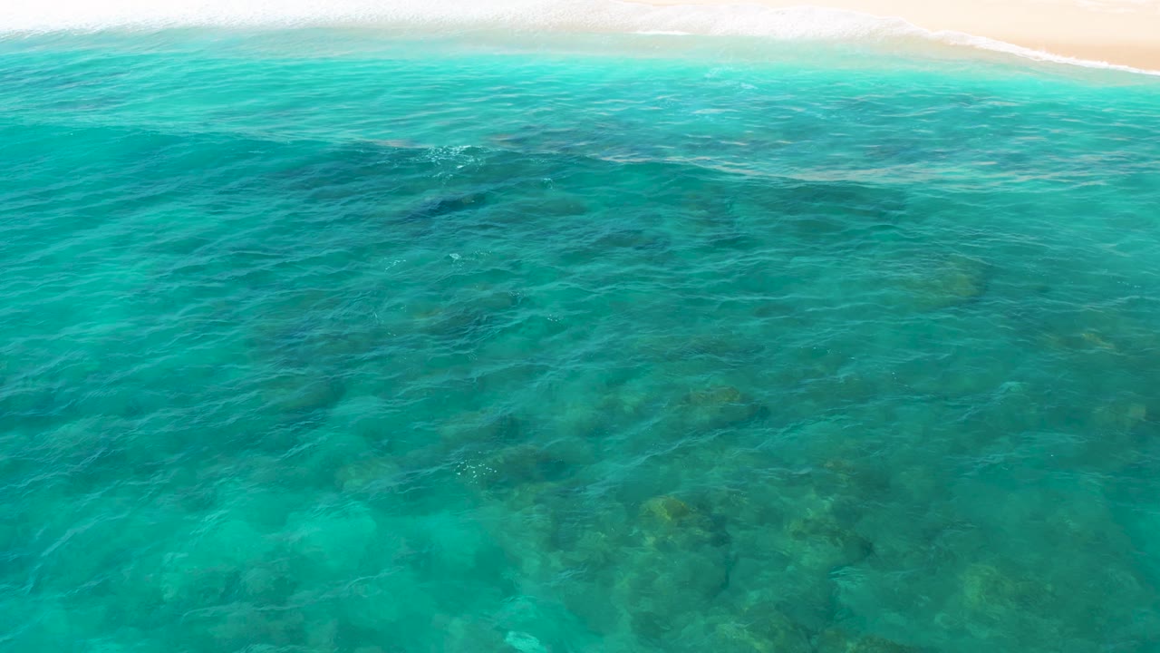 Blue-green ocean fills the aerial frame at El Cardoncito, with underwater rocks in the lower half and a narrow sandy shoreline with small breaking waves near the top, with no visible people.