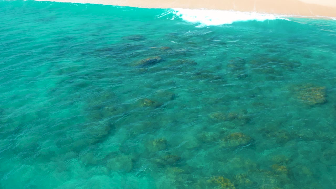 At 0:00, clear turquoise water in El Cardoncito reveals scattered submerged rocks, with a thin strip of sand and white wave wash along the top edge and no visible people.