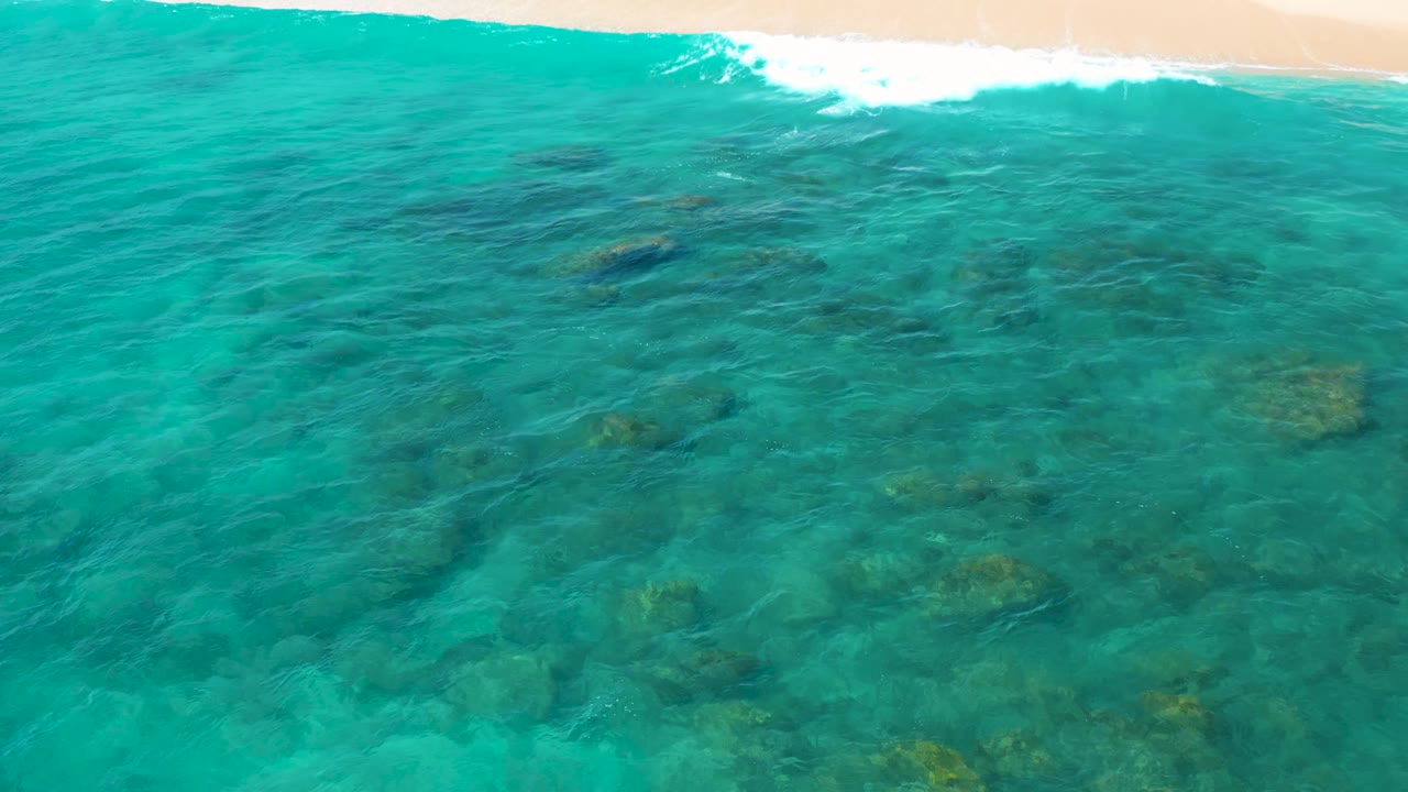 At 0:01, the view holds on shallow green-blue water in El Cardoncito, with darker submerged rock patches more centered and a small band of surf at the top, with no visible people.