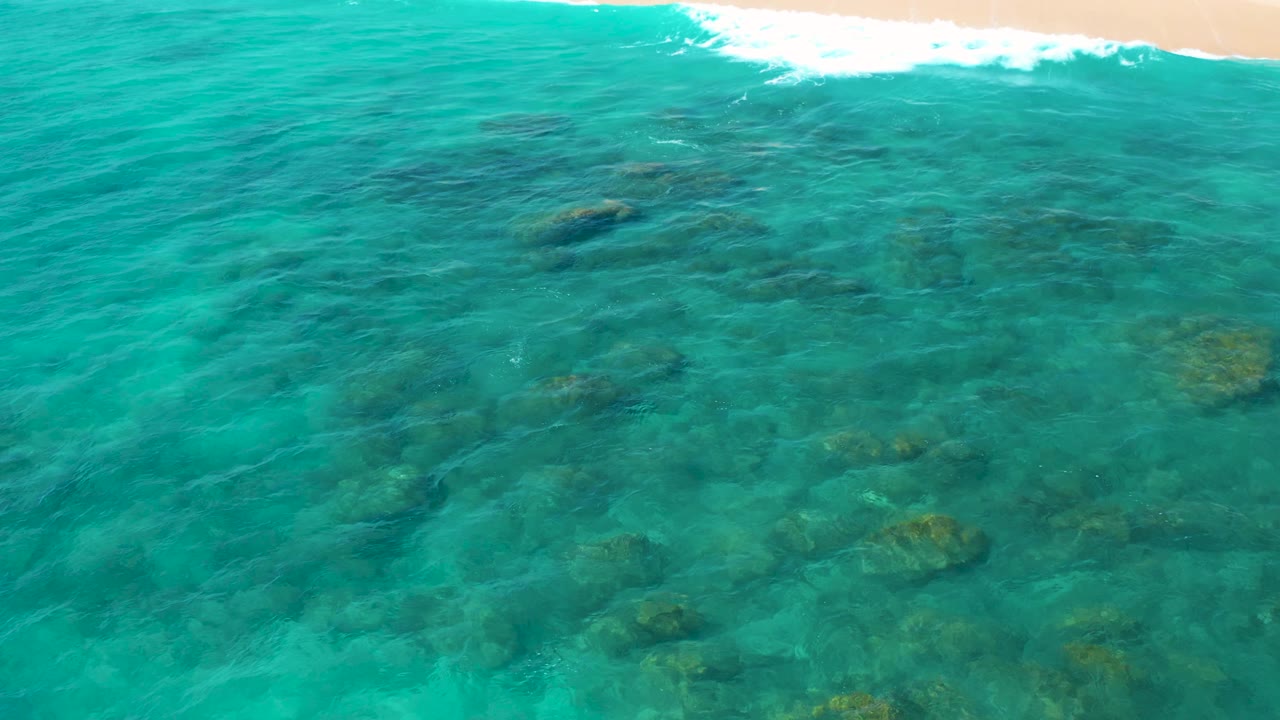 At 0:02, rippled turquoise water covers a rocky sea bottom in El Cardoncito, while pale sand and breaking white water remain visible along the upper frame, with no visible people.