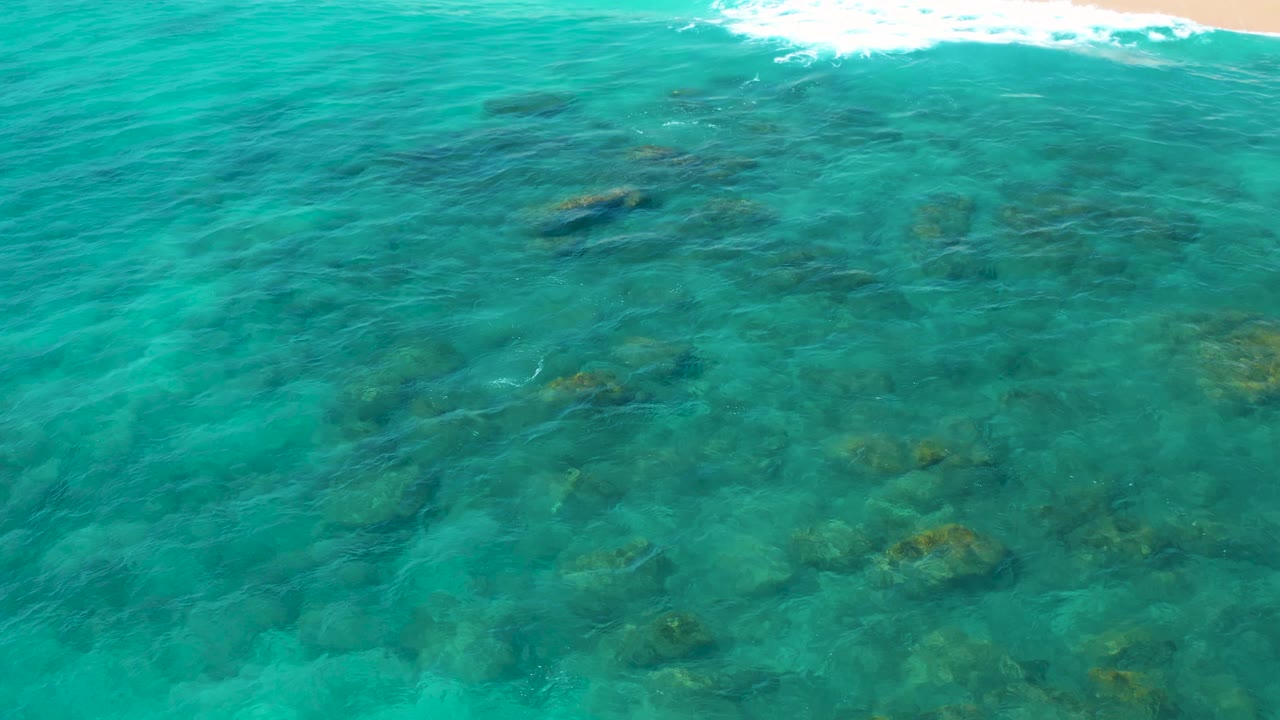 At 0:03, shallow green water in El Cardoncito fills most of the frame, with several darker rocks under the surface and a bright line of breaking water near the top, with no visible people.