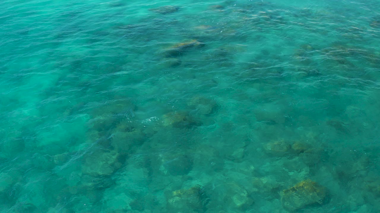 Overhead view of clear turquoise water over submerged rocks and sand patches in El Cardoncito, Baja California Sur, with no visible people.
