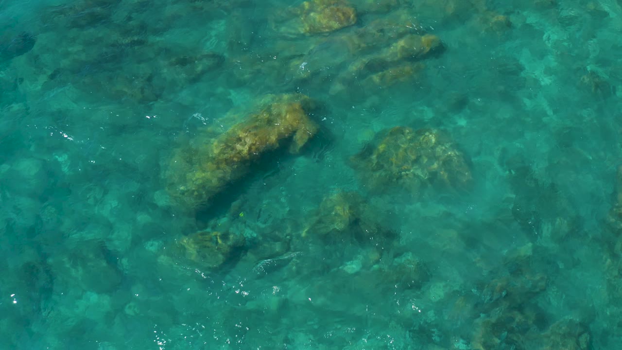 Aerial frame of rippled turquoise sea with several tan submerged rocks spread across the water at El Cardoncito, no visible people.