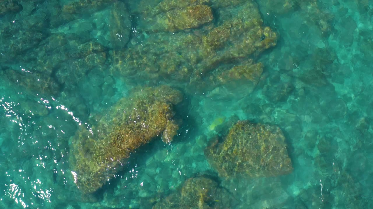 Clear blue-green water ripples over brown submerged rocks, with brighter reflected highlights near the lower left and no visible people.