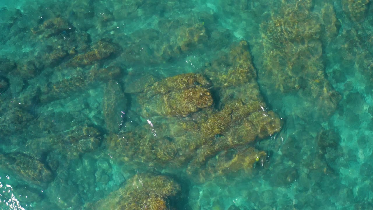 Blue-green shallow water covers large rounded rocks, with small white reflections tracing the moving surface and no visible people.