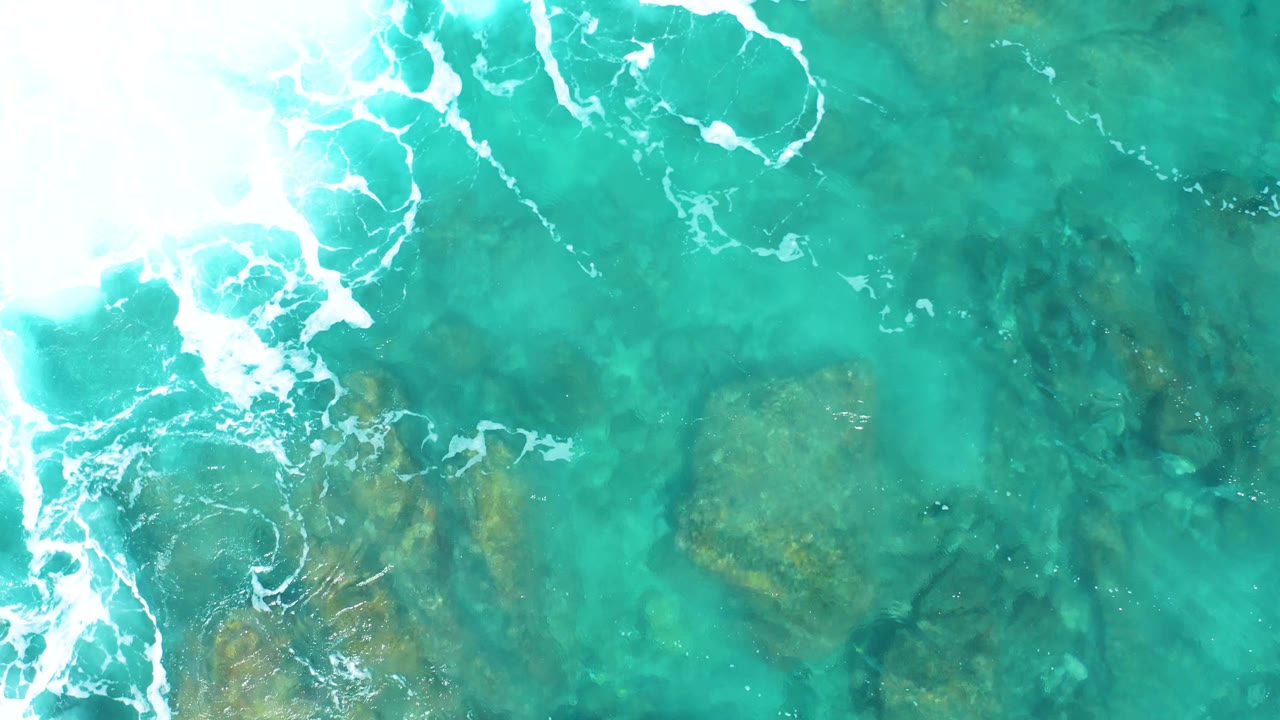The aerial composition shifts slightly over El Cardoncito water, with a larger foamy surf edge on the left and darker rocky patches under the green-blue surface to the right.