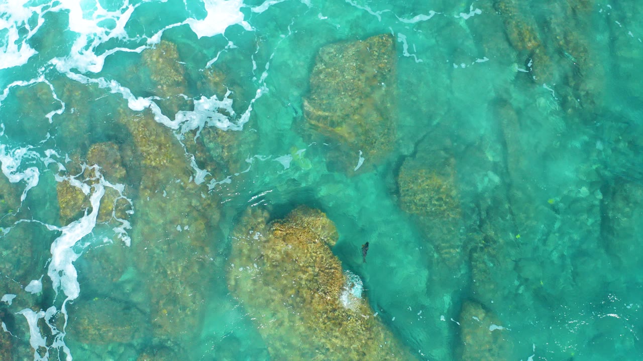 Overhead view of clear turquoise water washing around tan rock shelves at El Cardoncito, Baja California Sur, with white foam near the upper left and no visible people.