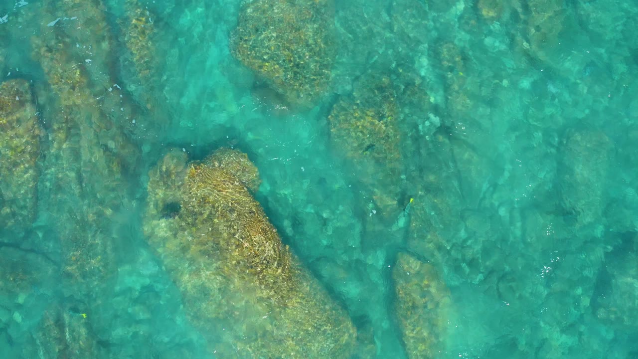 A top-down frame shows shallow turquoise sea and tan rock forms below the surface in El Cardoncito, Baja California Sur, with no visible people.