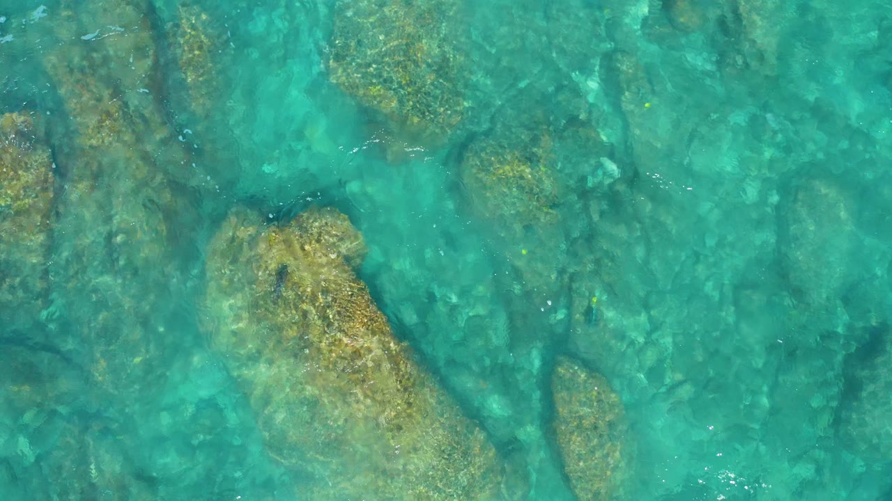 Turquoise water reveals a large tan submerged rock and darker underwater forms in El Cardoncito, Baja California Sur, with no visible people.