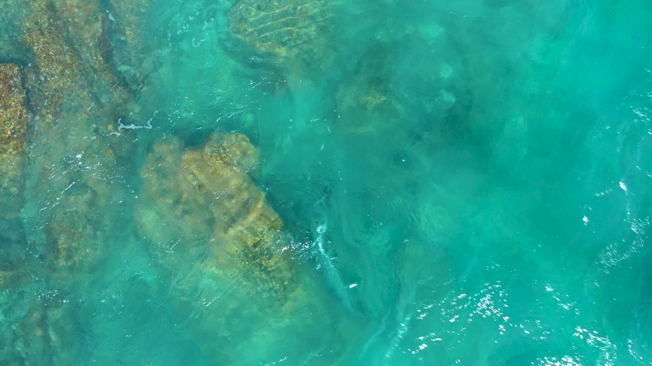 Top-down view of turquoise seawater over submerged rock shelves in El Cardoncito, Baja California Sur, with faint foam lines and no visible people.
