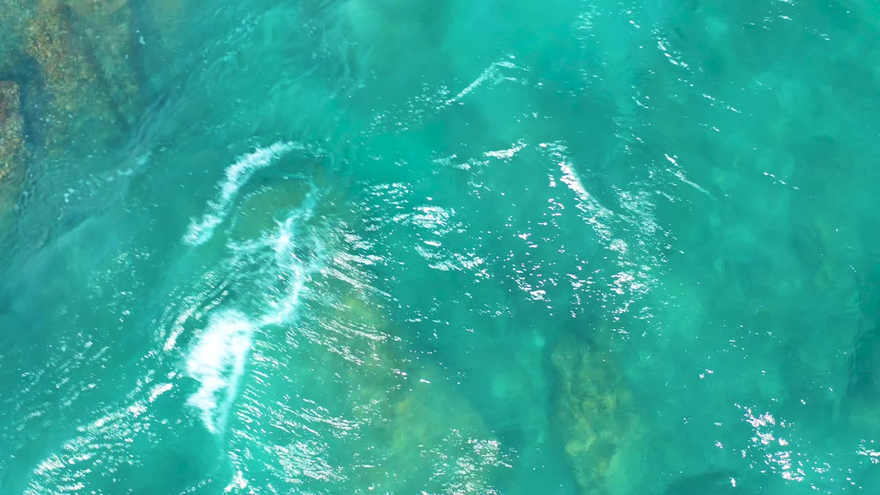 White foam curves across shallow turquoise water above submerged rocks in El Cardoncito, Baja California Sur, with no visible people.