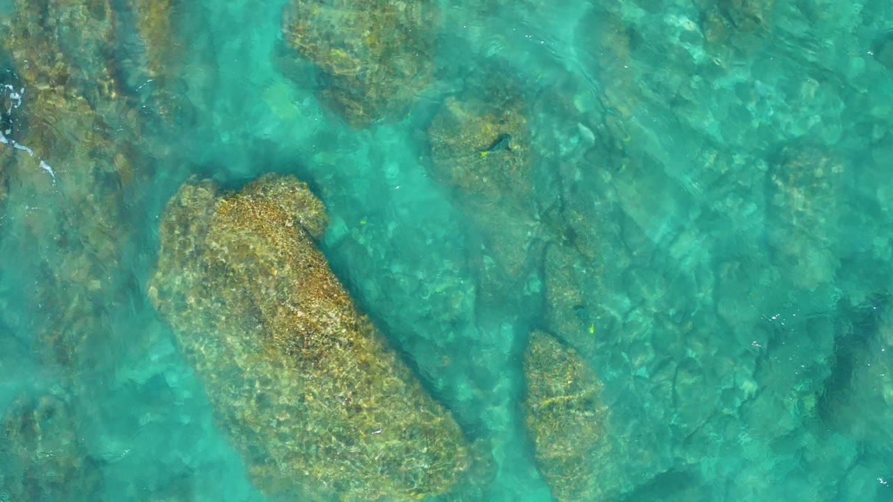 Clear turquoise shallows reveal a broad beige rock and smaller underwater formations in El Cardoncito, Baja California Sur, with no visible people.