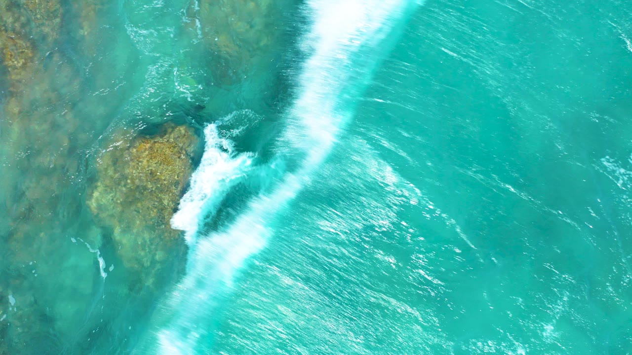 Overhead view at El Cardoncito, Baja California Sur, where white surf crosses turquoise water above tan submerged rocks, with no visible people.