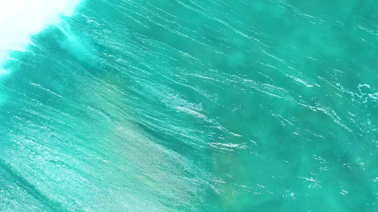 Turquoise water at El Cardoncito fills the overhead frame as the foam recedes toward the edge and sunlight reveals fine surface ripples, with no visible people.