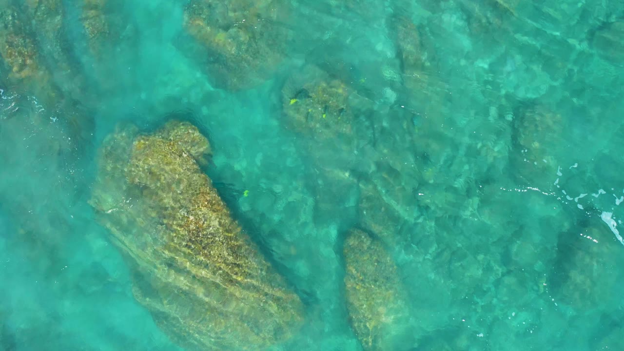 Overhead frame from El Cardoncito with blue-green water, a broad rock shape near center, smaller rocks nearby, and no visible people.