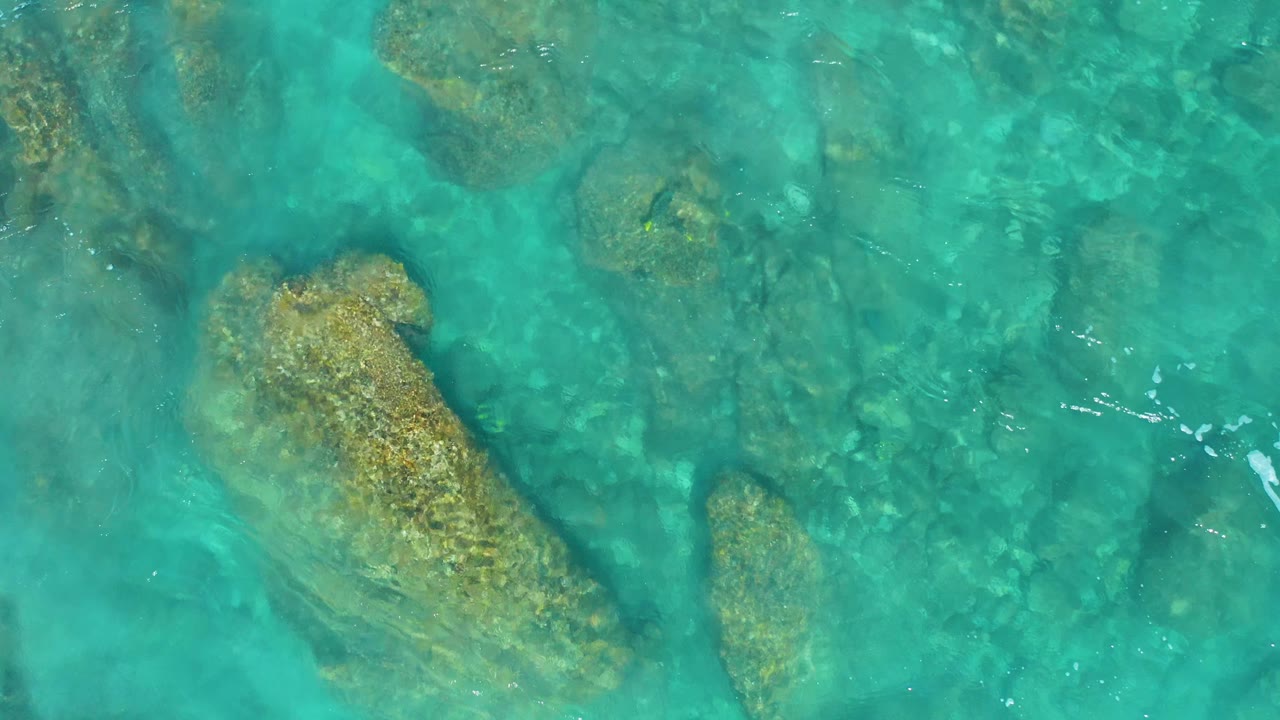 Overhead view of El Cardoncito rocky shallows, with a tan rock mass under bright turquoise water and no visible people.