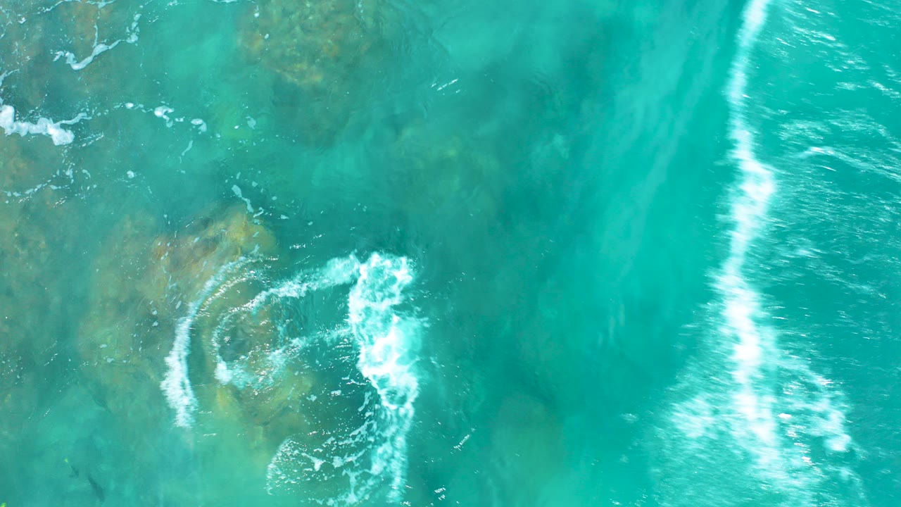 Overhead view of turquoise water at El Cardoncito, Baja California Sur, with white wave foam curling over shallow submerged rocks and no visible people.