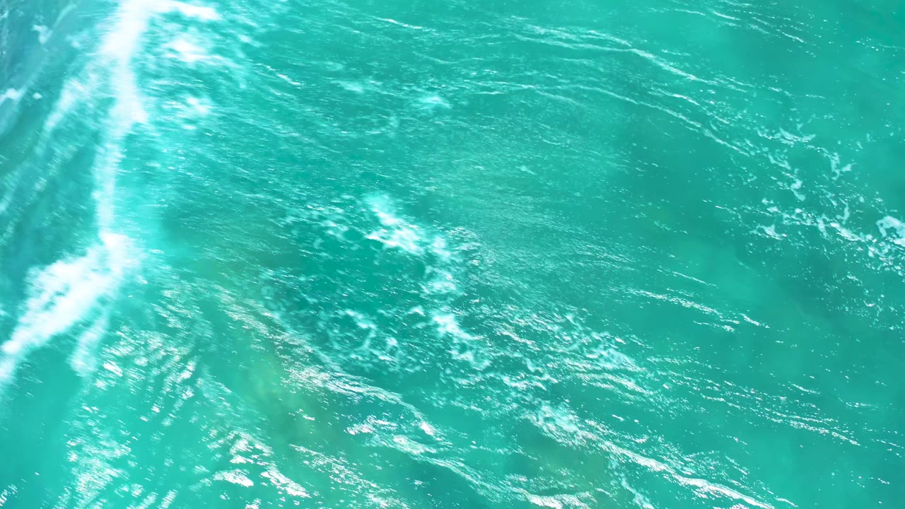 Top-down El Cardoncito water view with bright white wave streaks crossing the turquoise surface and darker seabed tones faintly visible below, with no visible people.