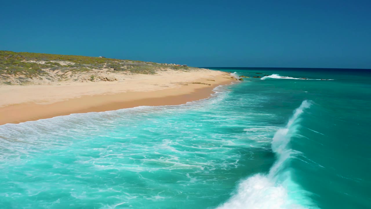 Aerial frame from El Cardoncito showing a white wave line rolling diagonally toward the sand, with low dunes behind the beach and no visible people.