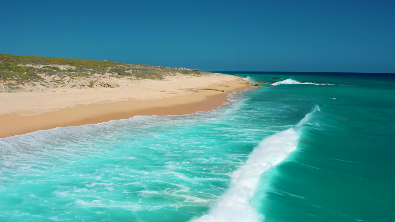 Aerial view at El Cardoncito with a larger breaking wave crest near the right side of frame, open blue ocean beyond, sandy beach, and no visible people.