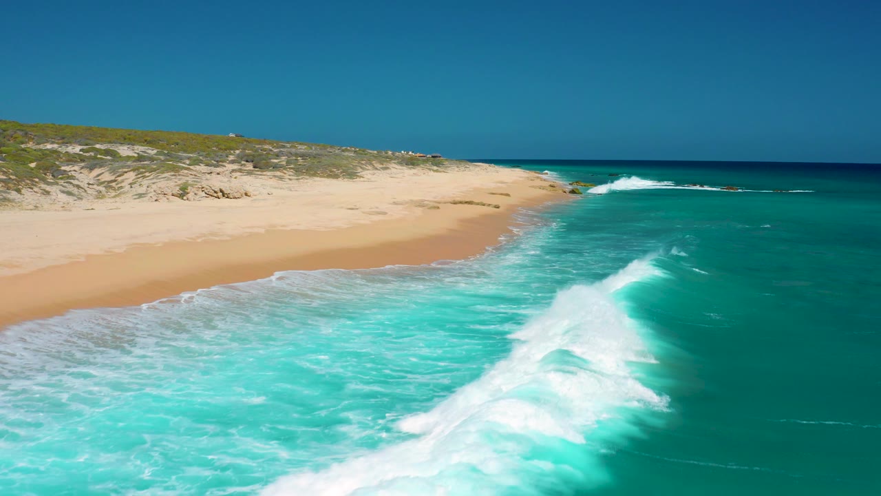 Foamy surf spreads across turquoise water near the empty El Cardoncito beach, with pale sand and low coastal vegetation visible beneath a clear sky.