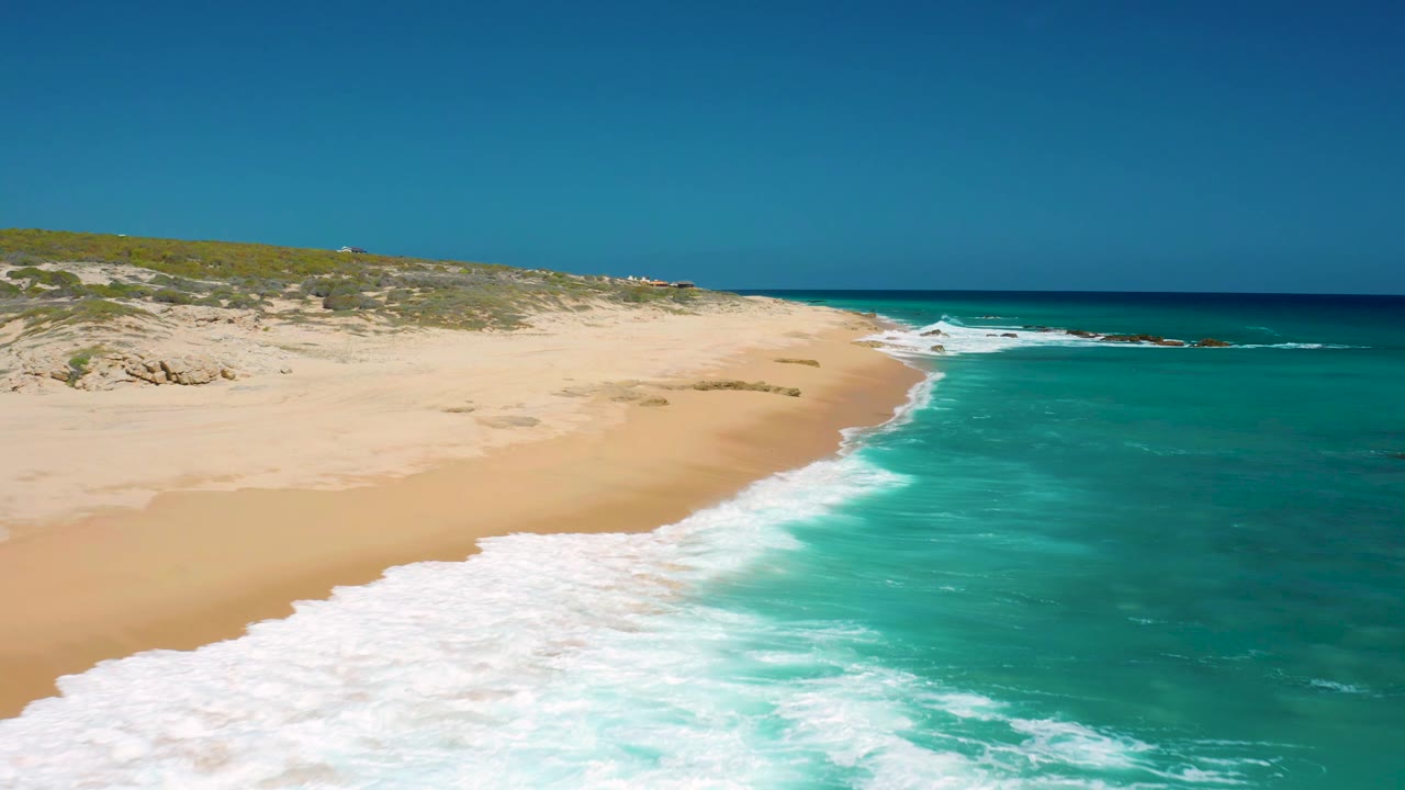 Aerial frame from El Cardoncito showing a broad band of foam moving up the beach, clear blue-green water to the right, low brush beyond the sand, and no visible people.