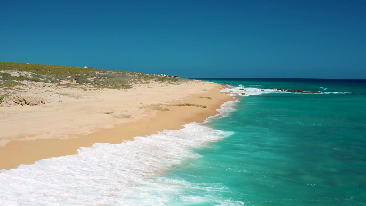 Aerial view over El Cardoncito with turquoise water occupying the right side, an empty sandy beach on the left, low vegetation behind the beach, and no visible people.