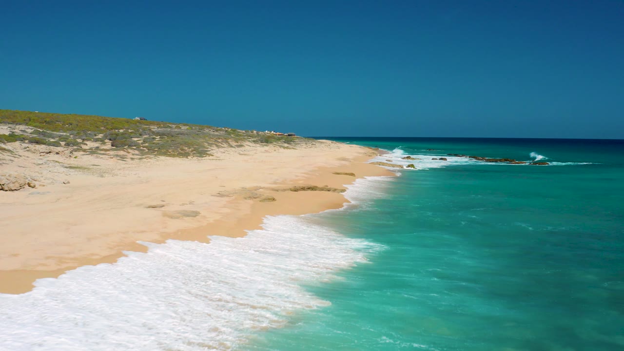 Aerial frame at El Cardoncito where the foamy surf cuts diagonally across the lower left, the sandy beach opens toward the dunes, turquoise water sits to the right, and no visible people appear.