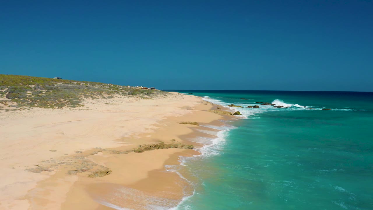 A second aerial view of El Cardoncito shows the shoreline extending diagonally, white foam tracing the wet sand, and no visible people on the beach.