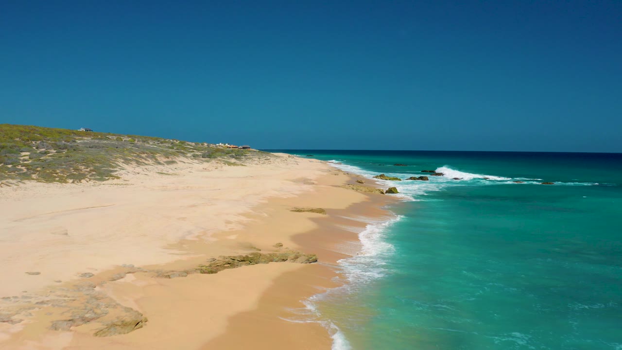 The aerial perspective over El Cardoncito moves along an empty sandy shore, with bright turquoise water, white breaking waves, small rocks near the surf, and no visible people.