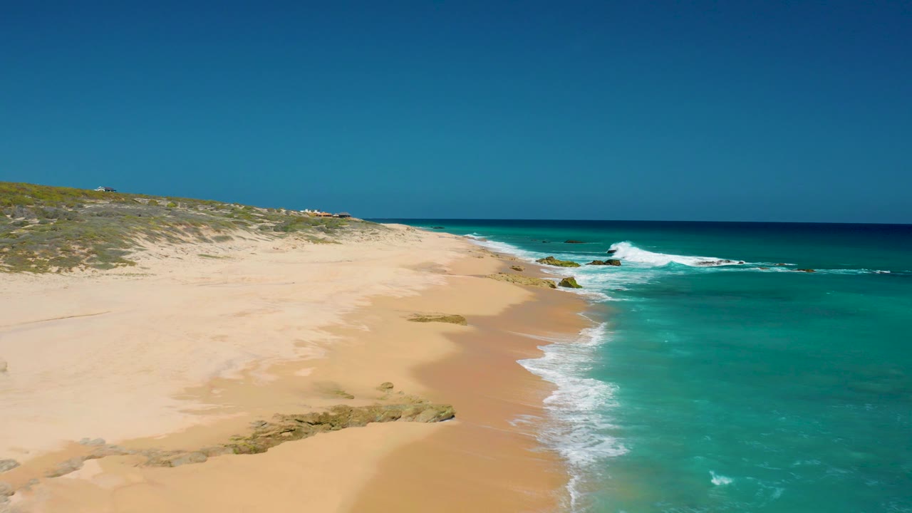 Mid-clip aerial frame in El Cardoncito showing a broad sandy foreground, foamy surf wrapping around rocks, a straight blue horizon, and no visible people.