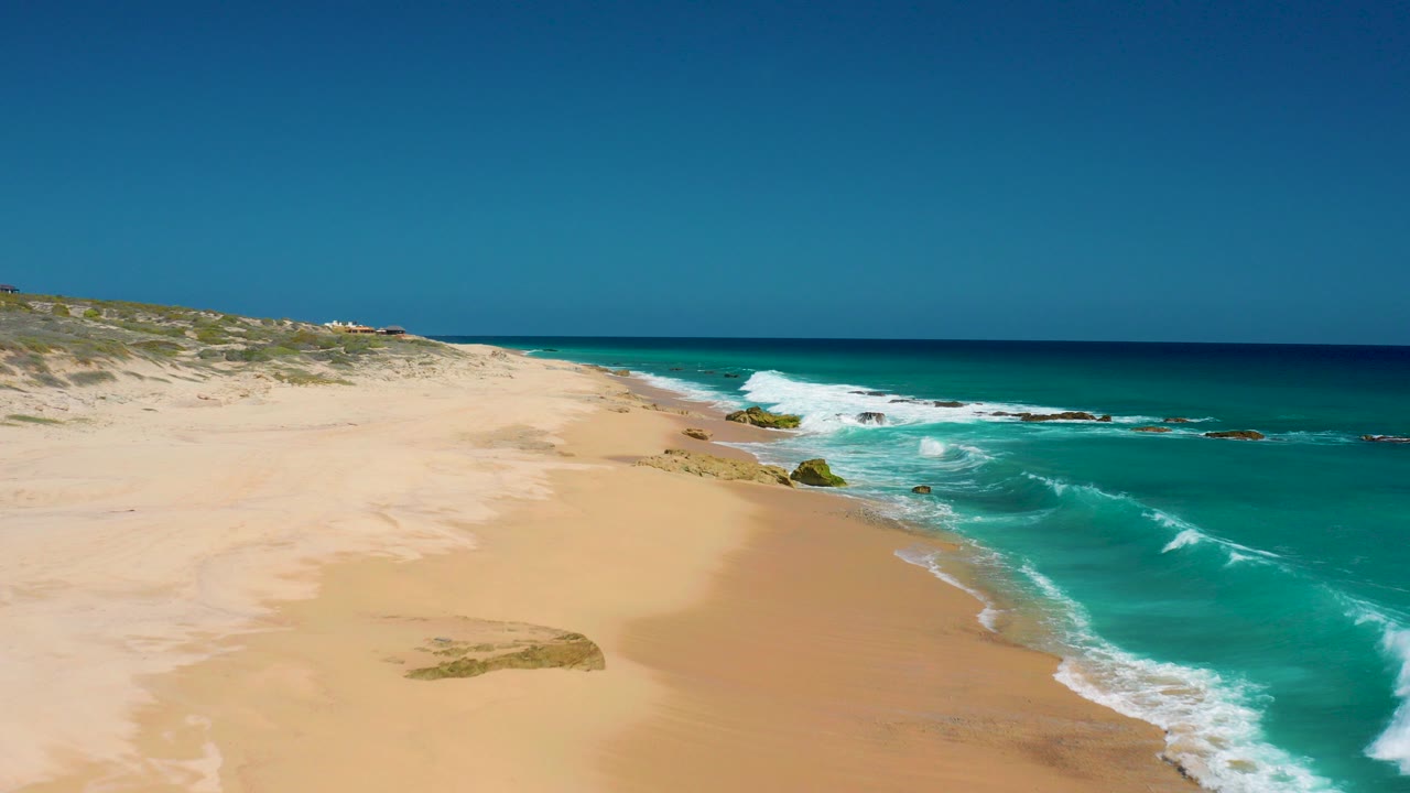 Wide aerial view at El Cardoncito, Baja California Sur, with empty sand on the left, turquoise ocean on the right, low dunes beyond the beach, and no visible people.